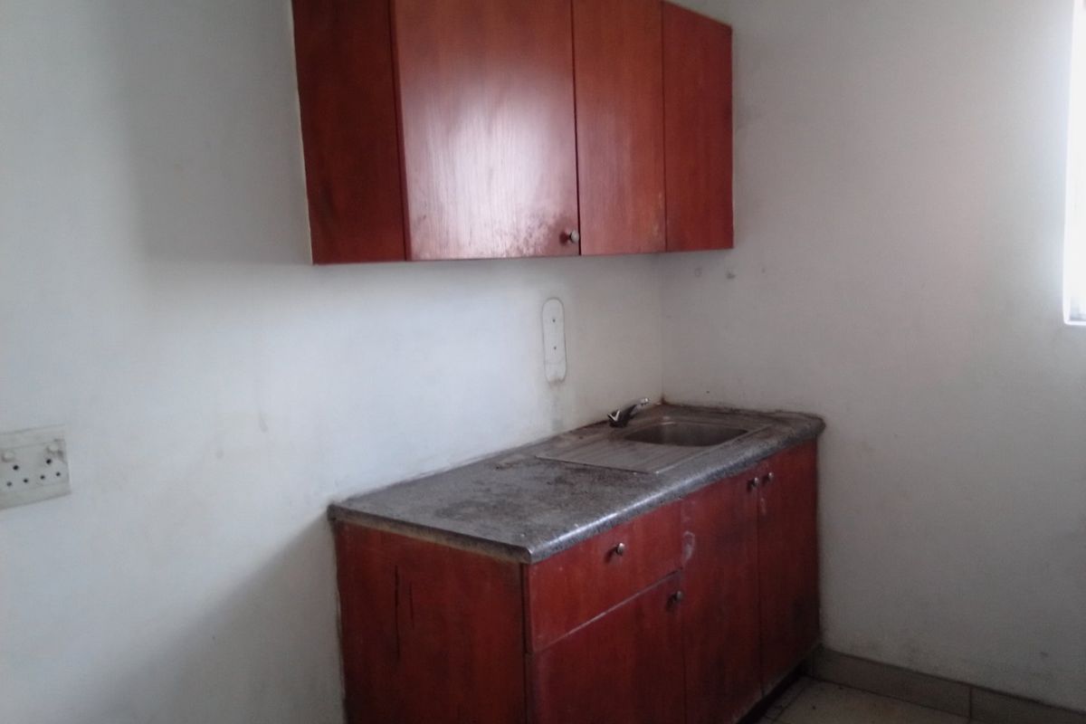 A compact kitchen area featuring built-in wooden cabinetry, a stainless steel sink, and tiled flooring, currently in need of cosmetic refurbishment.
