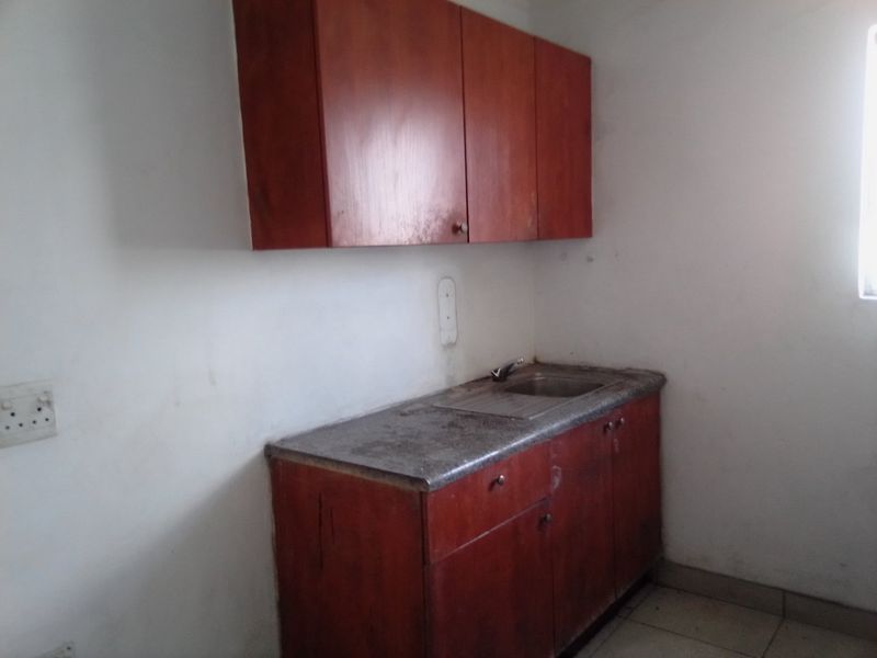 A compact kitchen area featuring built-in wooden cabinetry, a stainless steel sink, and tiled flooring, currently in need of cosmetic refurbishment.