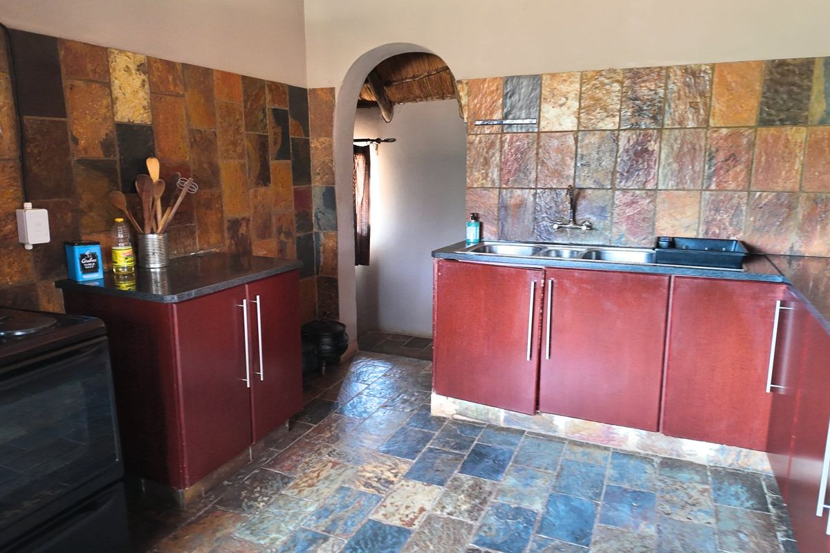 A rustic kitchen featuring stone tile flooring and backsplash, dark cabinetry, and a double basin sink, connected to an adjacent space via an archway.