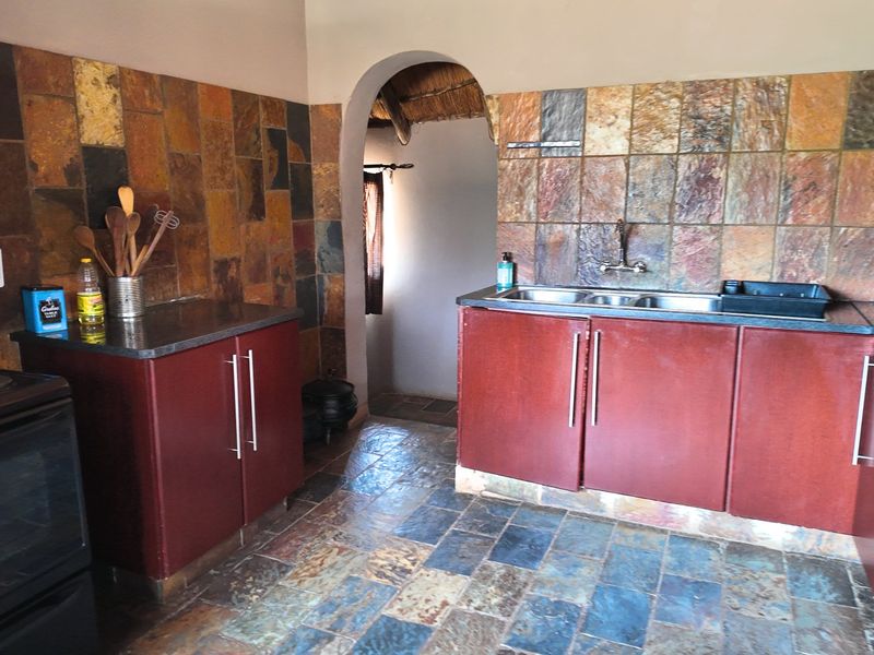 A rustic kitchen featuring stone tile flooring and backsplash, dark cabinetry, and a double basin sink, connected to an adjacent space via an archway.