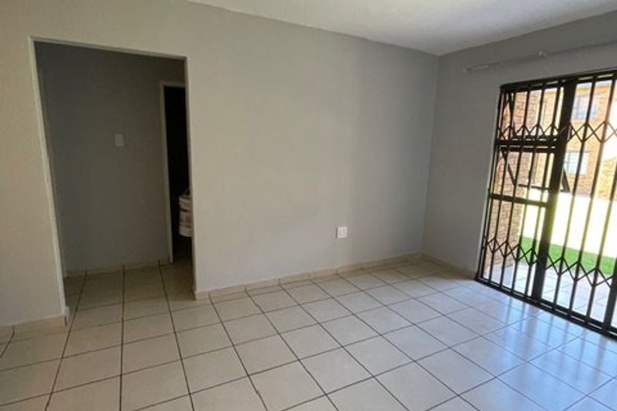 An empty, neutrally painted living room featuring ceramic floor tiles and a security gate leading to the exterior.
