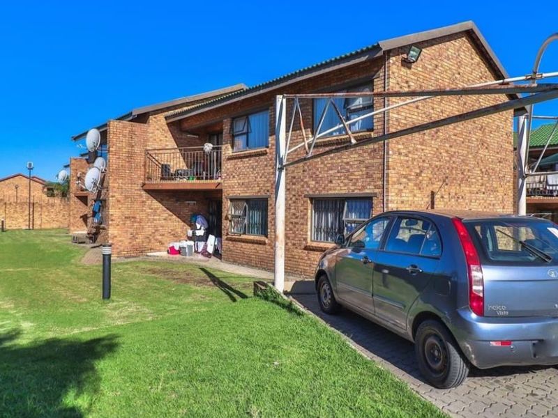 Rear view of a face brick apartment building featuring a balcony, paved driveway, and a shared lawn area.