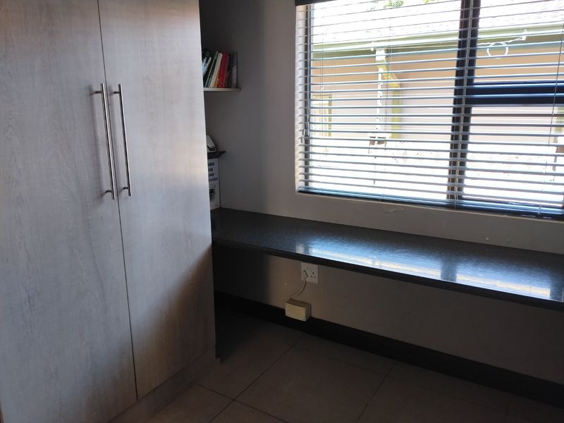 A functional home office space featuring a built-in desk, ample cabinetry, tiled flooring, and large windows with fitted blinds.