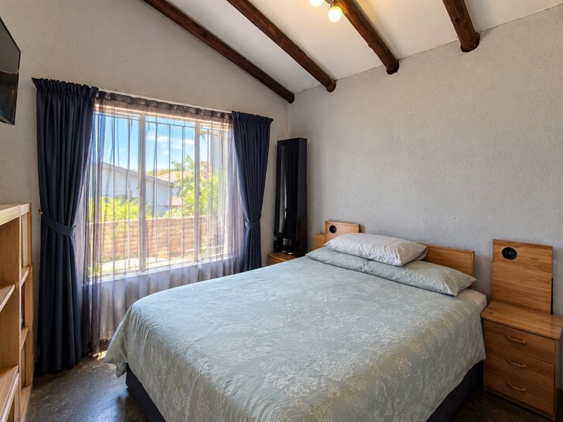 A bedroom featuring exposed timber ceiling beams, built-in headboard and bedside pedestals, and a window with security bars.