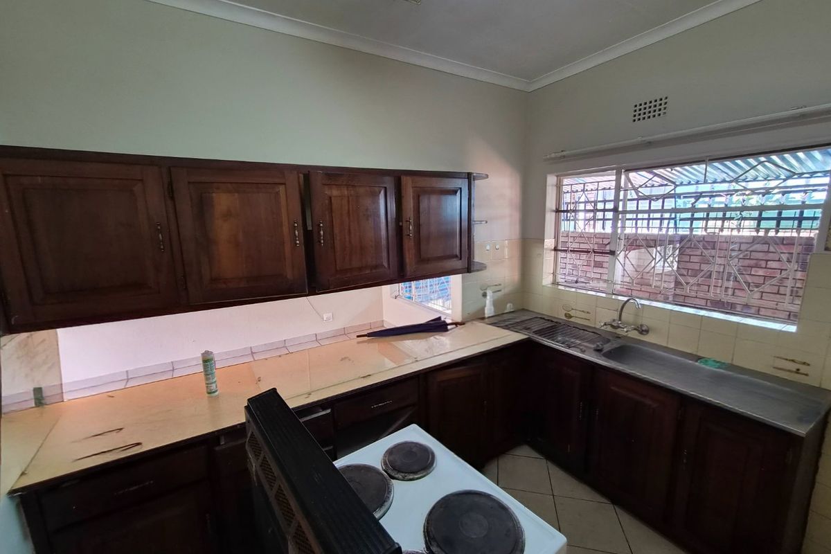 A dated kitchen featuring dark wooden cabinetry, laminate countertops, a stainless steel sink, and an electric stove, with security bars on the window.