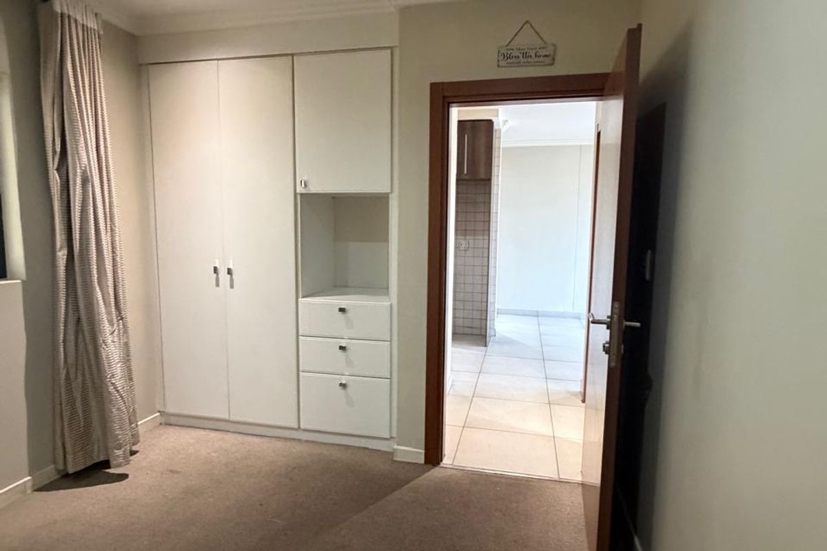 A clean, empty bedroom featuring built-in white cupboards, carpeted flooring, and recessed lighting, with a doorway leading to an adjacent tiled living area.