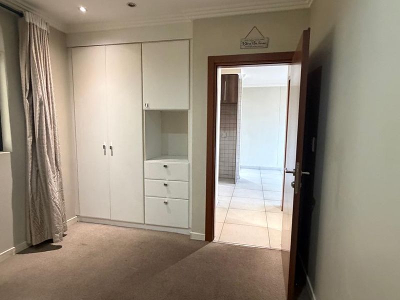 A clean, empty bedroom featuring built-in white cupboards, carpeted flooring, and recessed lighting, with a doorway leading to an adjacent tiled living area.
