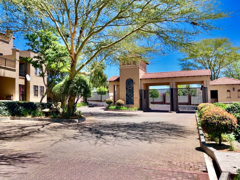 A well-maintained apartment complex entrance featuring a secure gated access point, paved driveway, and established landscaped gardens.