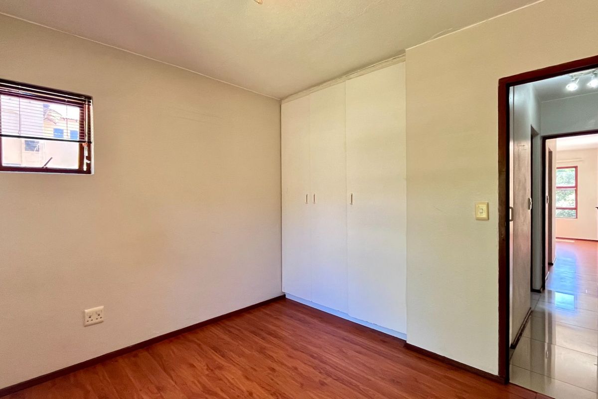A clean, empty bedroom featuring laminate flooring, built-in cupboards, and a window with blinds, offering a neutral space ready for personalization.