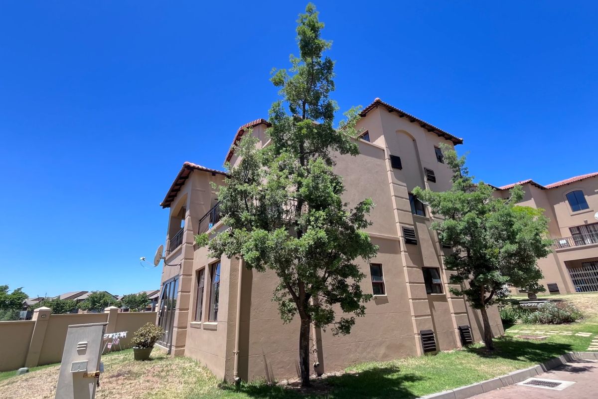 Exterior view of a multi-story apartment building featuring a stucco facade, terracotta roof tiles, and a balcony, set within a landscaped residential complex.