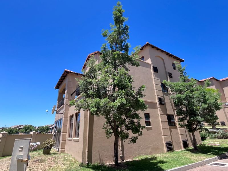 Exterior view of a multi-story apartment building featuring a stucco facade, terracotta roof tiles, and a balcony, set within a landscaped residential complex.
