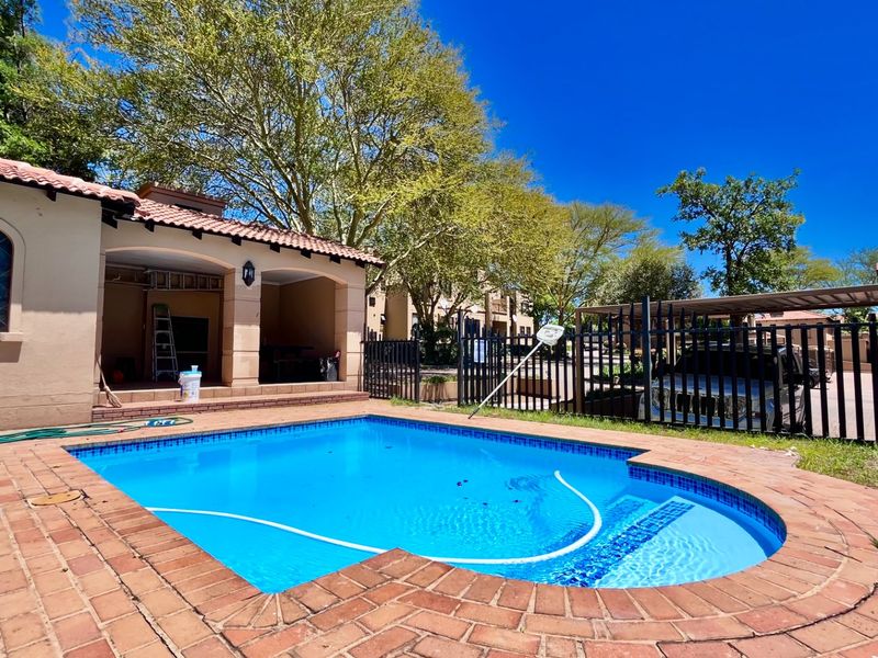 Outdoor pool area featuring a swimming pool with brick coping, adjacent covered patio, and secure metal perimeter fencing.