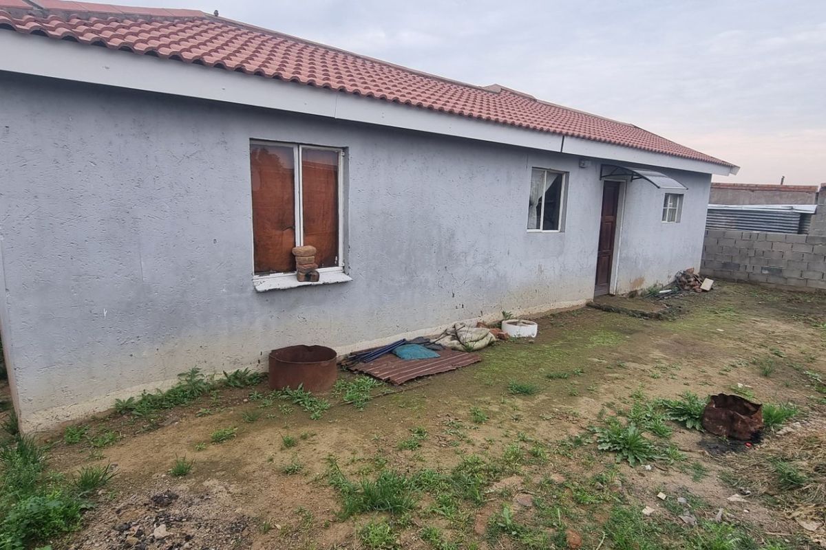 Rear view of a single-story house featuring a tiled roof and plastered exterior walls, set on an unlandscaped plot with a concrete block boundary wall.