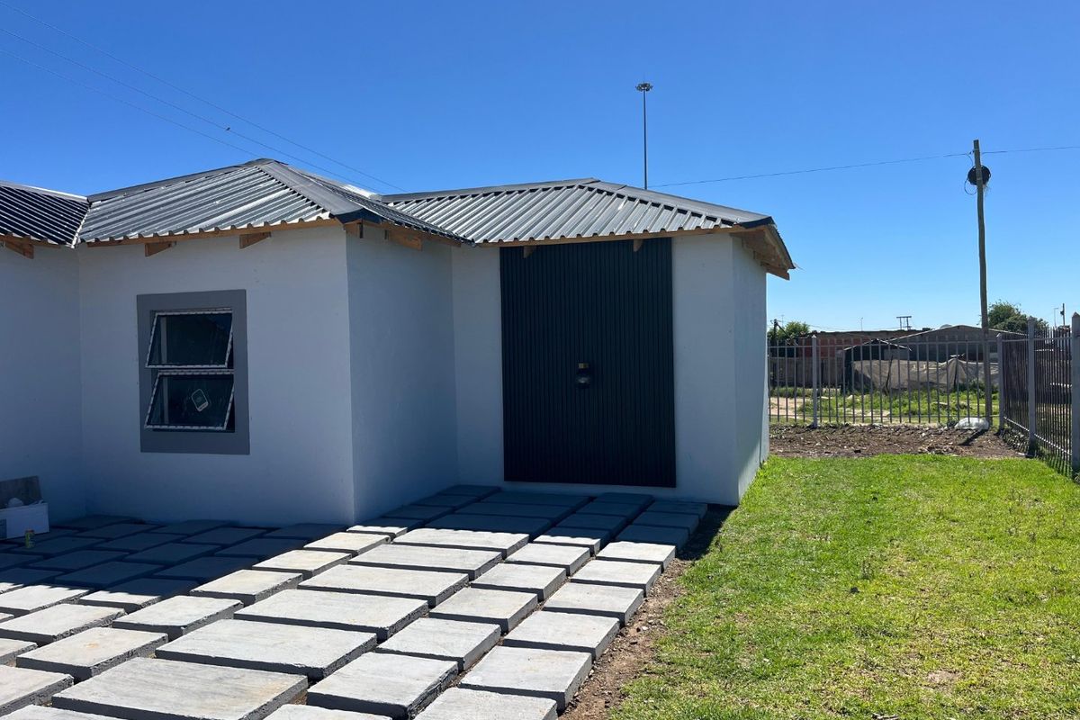 Rear view of a modern house featuring a corrugated metal roof, paved walkway, and secure metal door, set within a fenced yard.