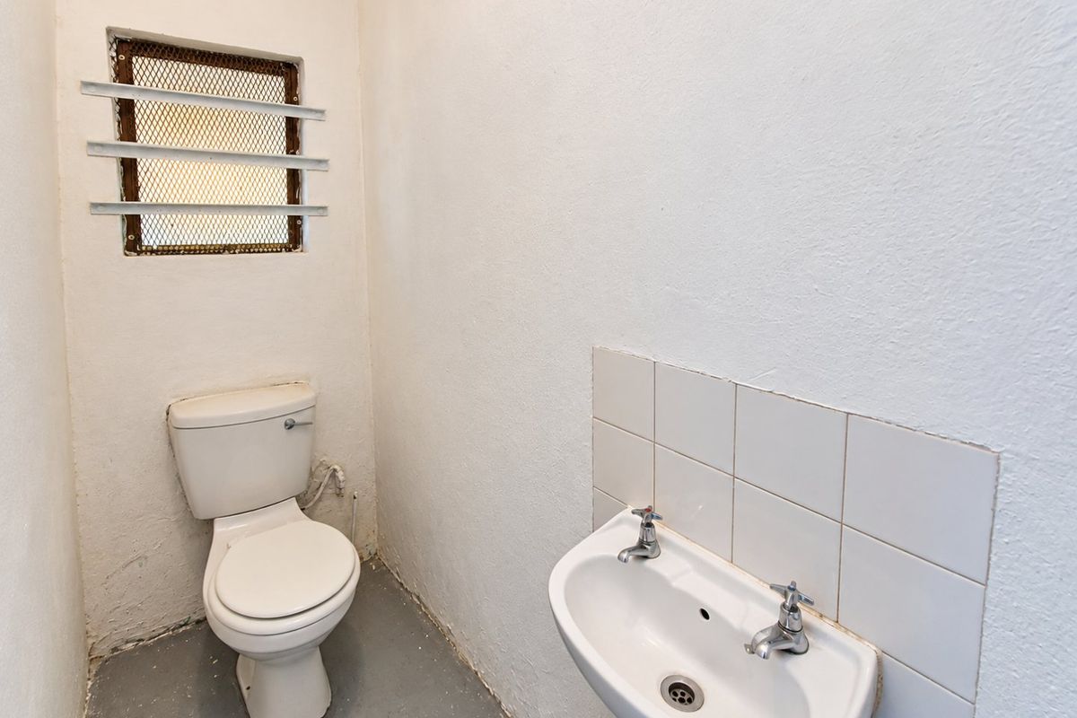 A basic, functional bathroom featuring a wall-mounted basin with tiled splashback, a standard toilet, and a small window with security bars.