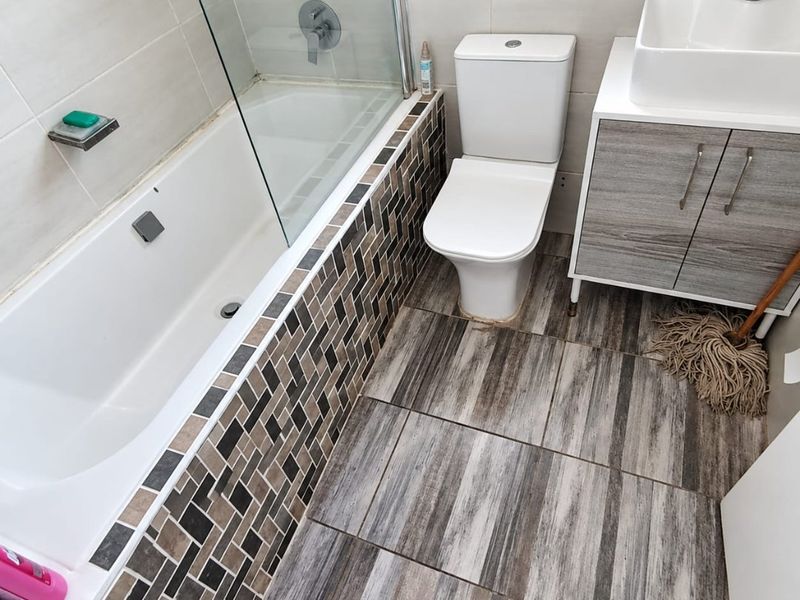 A compact bathroom featuring a bathtub with a glass screen, a modern vanity with basin, and a toilet, finished with wood-look floor tiles.
