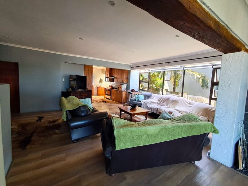 Spacious open-plan living area featuring laminate flooring, exposed timber beams, and large windows, seamlessly flowing into a functional kitchen space.
