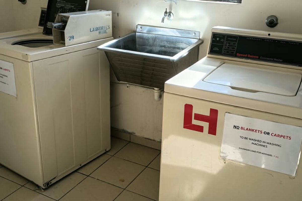A functional communal laundry area featuring tiled flooring, a stainless steel utility sink, and commercial-style washing machines.