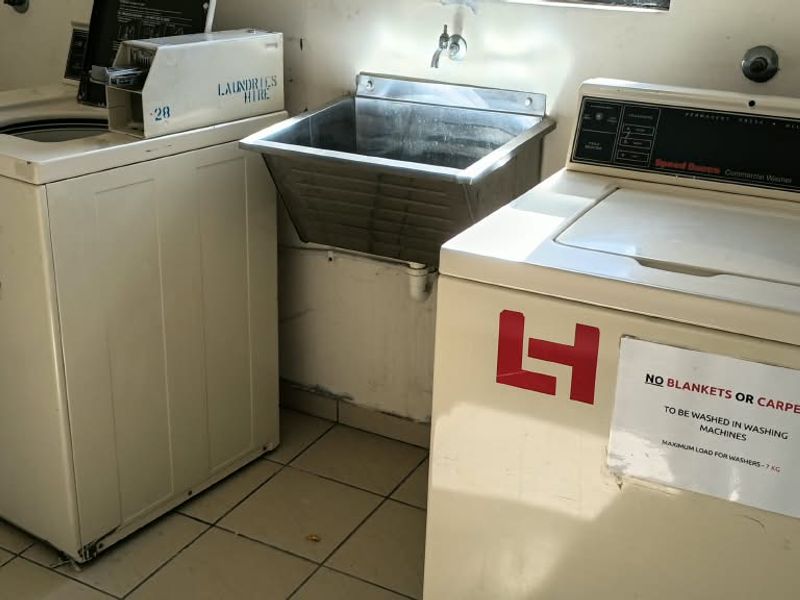 A functional communal laundry area featuring tiled flooring, a stainless steel utility sink, and commercial-style washing machines.