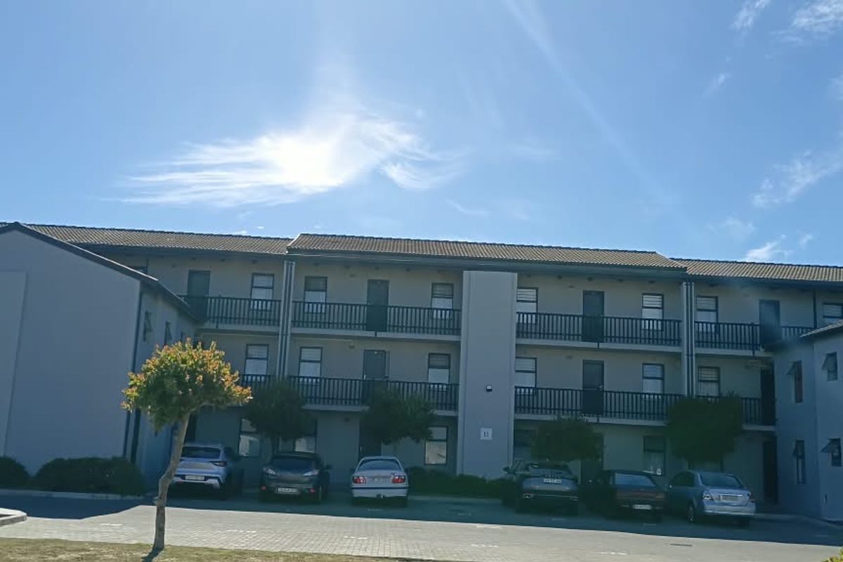 Front elevation of a multi-story apartment complex featuring balconies with railings and an adjacent paved parking area.