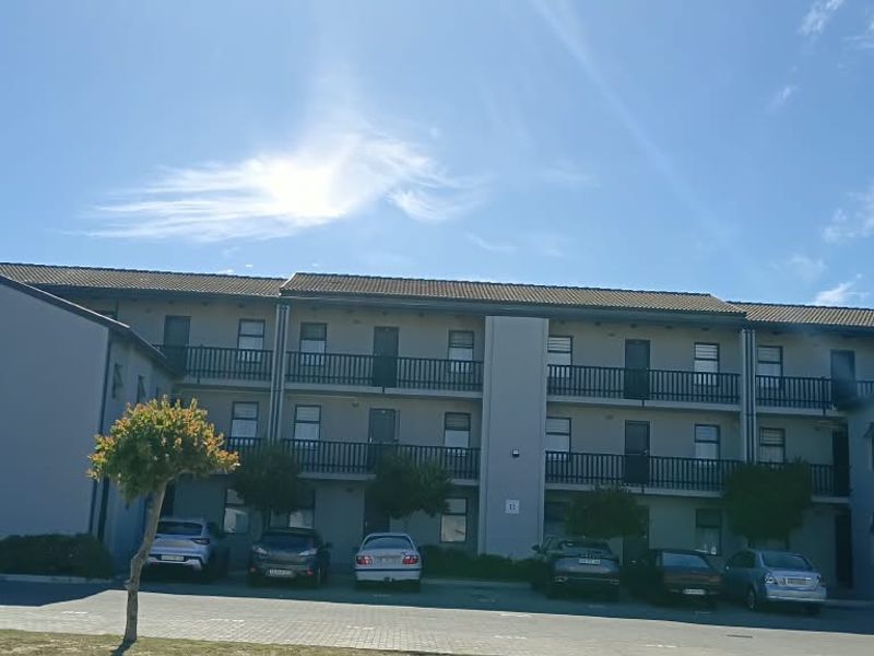 Front elevation of a multi-story apartment complex featuring balconies with railings and an adjacent paved parking area.