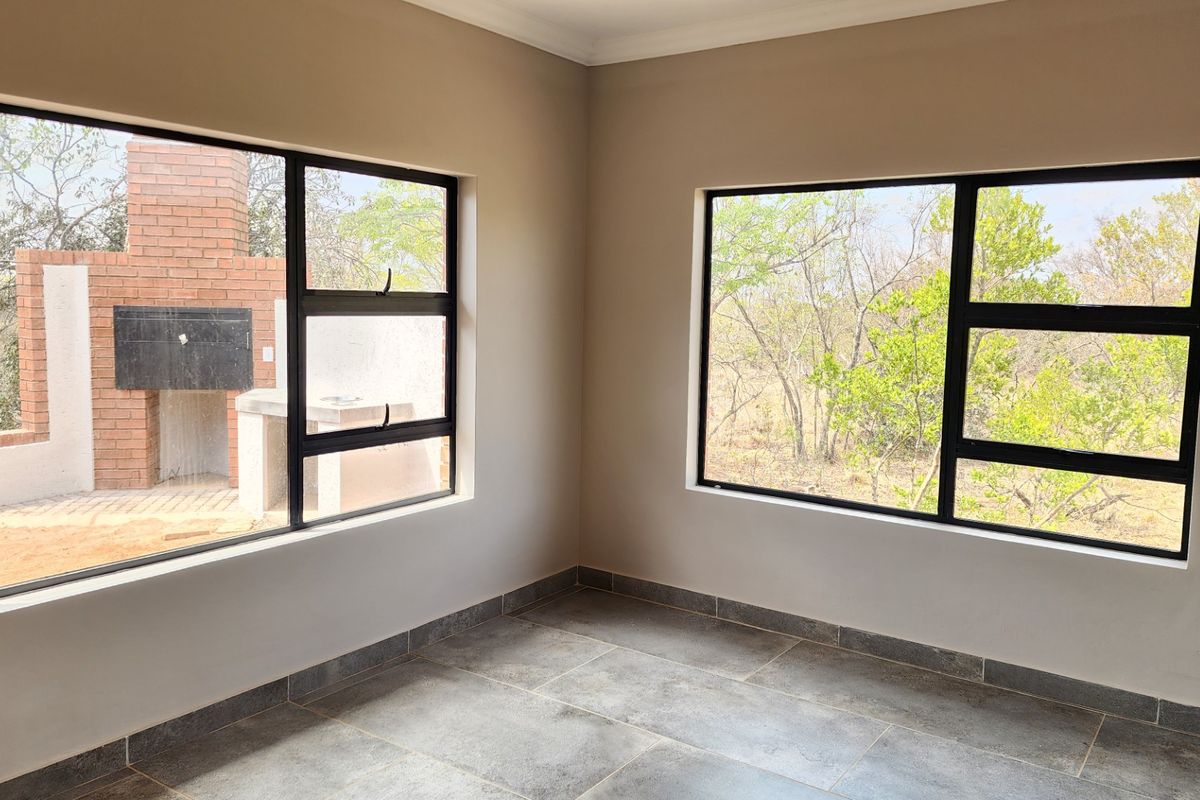 A bright, empty living area featuring large aluminium-framed windows with views of the outdoor brick braai area and natural surroundings.