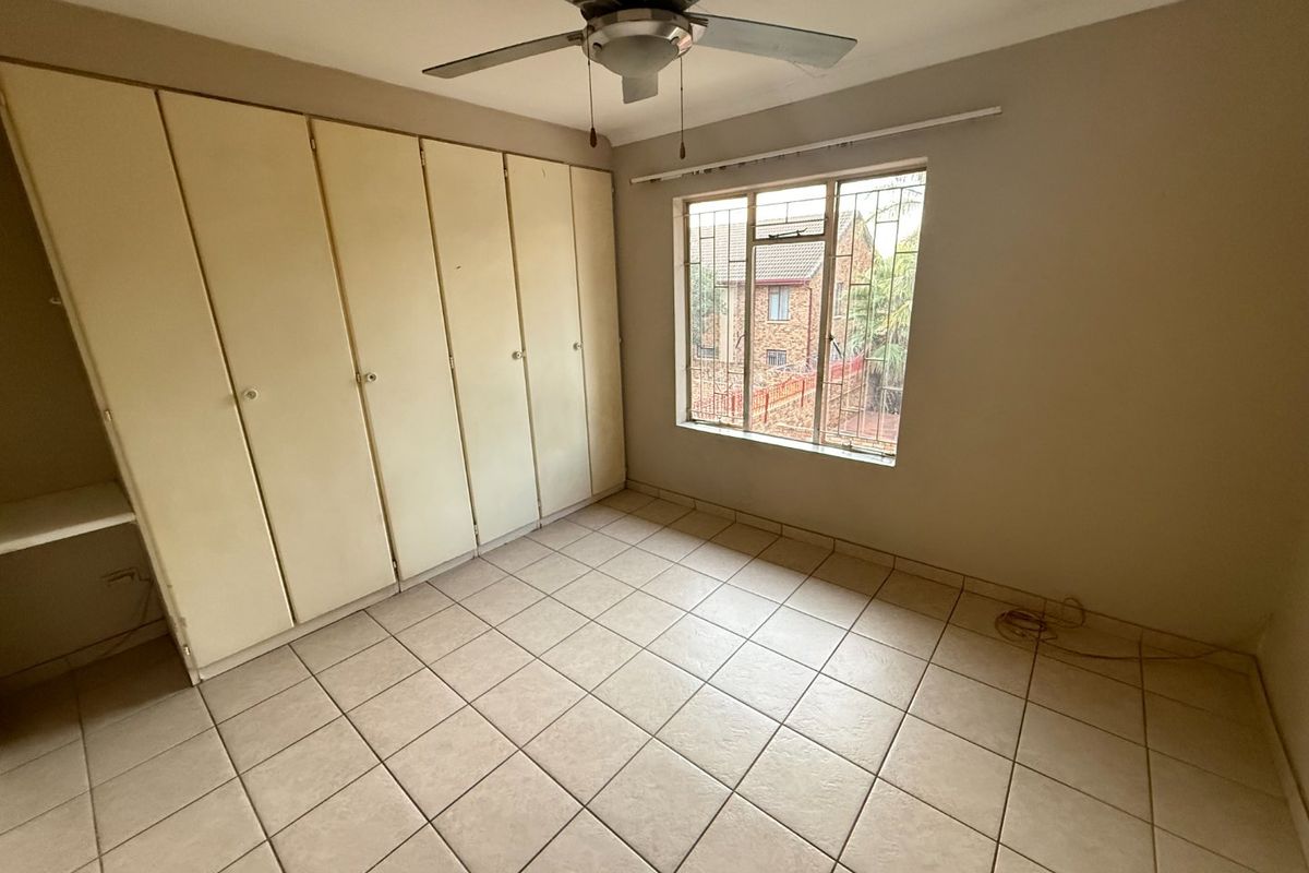 A spacious bedroom featuring neutral tiled flooring, extensive built-in cupboards, a ceiling fan, and a window fitted with security bars.