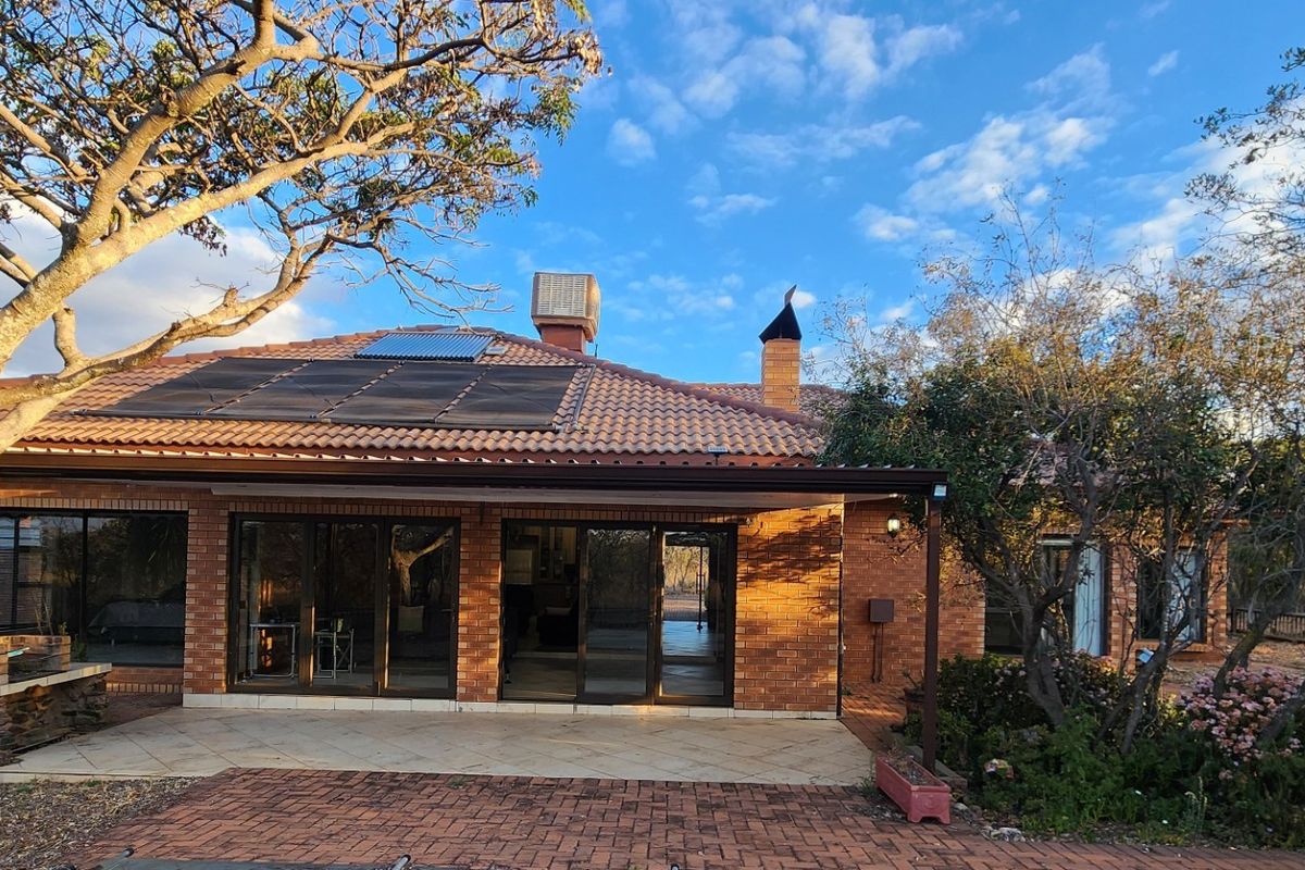 Rear view of a brick house featuring a covered patio, built-in braai, sliding glass doors, and solar water heating panels on a tiled roof.