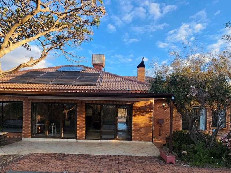 Rear view of a brick house featuring a covered patio, built-in braai, sliding glass doors, and solar water heating panels on a tiled roof.
