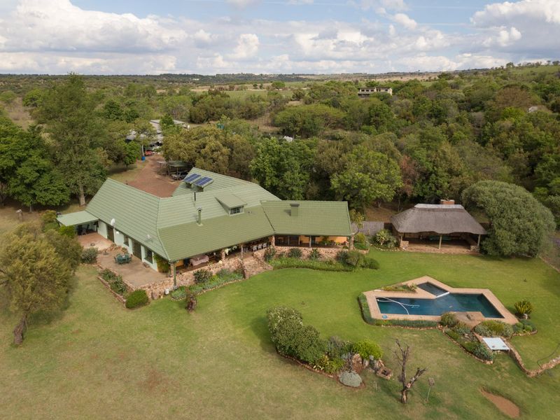 Aerial view of a spacious property featuring a large house with a green tiled roof, solar panels, a swimming pool, and a separate thatched lapa.
