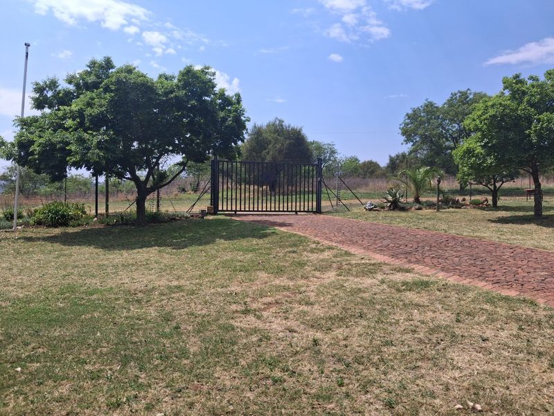 A view of the property entrance featuring a secure metal gate, a brick-paved driveway, and established garden landscaping.