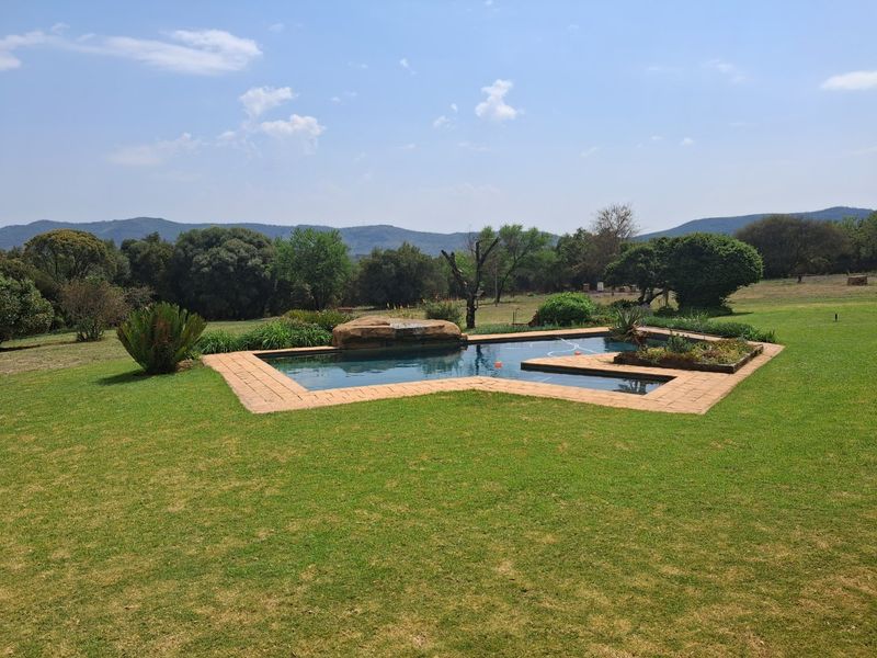 A well-maintained swimming pool featuring a natural rock water feature, surrounded by a manicured lawn and established garden with scenic mountain views.