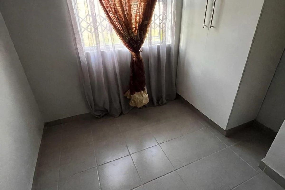 A compact bedroom featuring neutral tiled flooring, built-in cupboards, and a window fitted with a security gate.