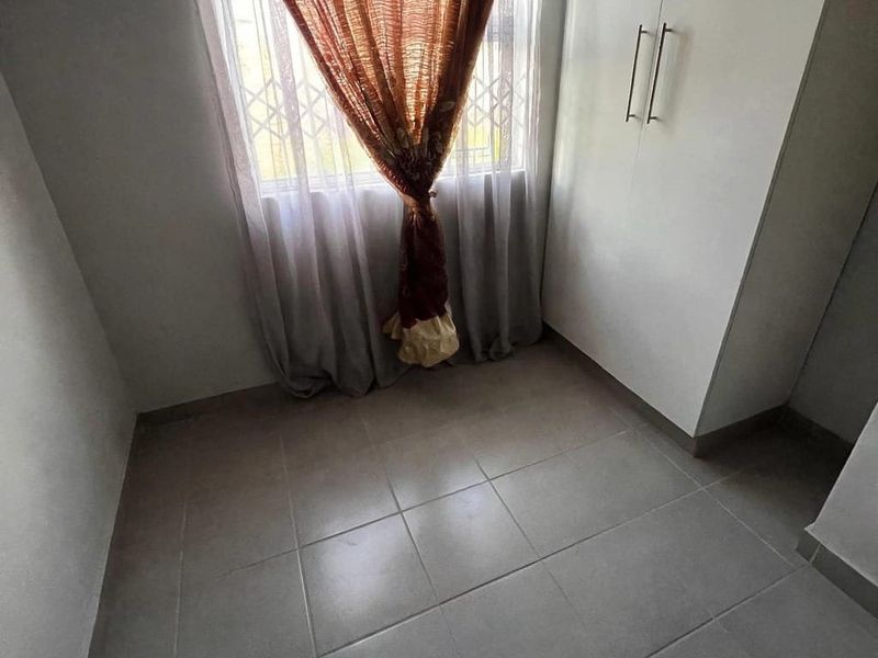 A compact bedroom featuring neutral tiled flooring, built-in cupboards, and a window fitted with a security gate.