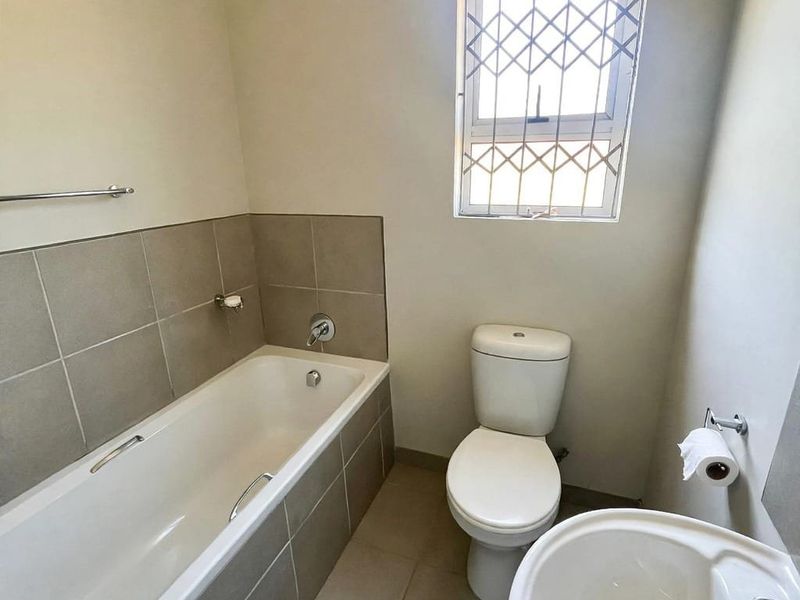 A clean, functional bathroom featuring a bathtub with tiled surround, pedestal basin, and toilet, finished with neutral wall tones and a security-fitted window.