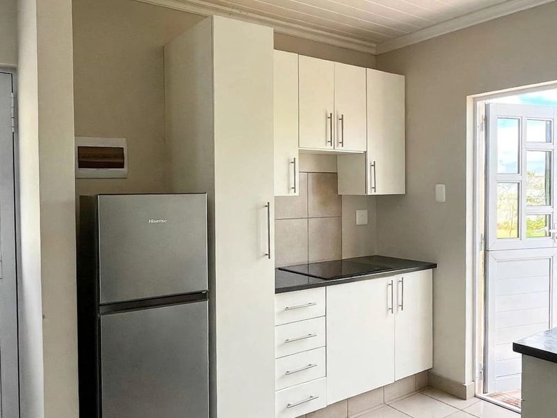 A compact kitchen featuring white built-in cabinetry, a black hob, tiled backsplash, and tiled flooring with direct access to an outdoor area.