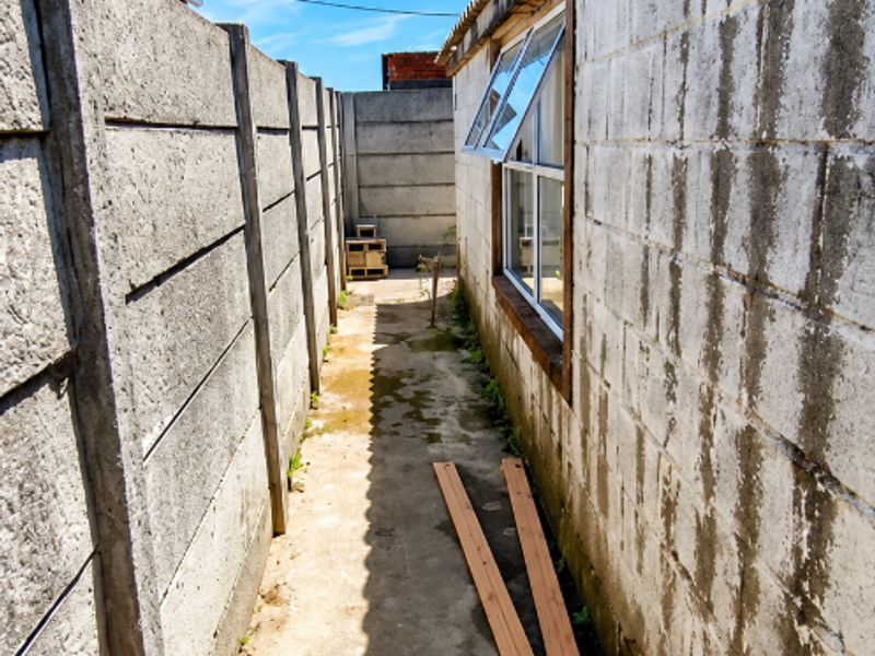 A narrow side passage featuring a concrete pathway, precast boundary wall, and exterior masonry wall with drainage infrastructure.
