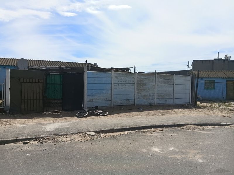 Street view of a property featuring a precast concrete boundary wall and metal security gate with corrugated roof structures visible in the background.