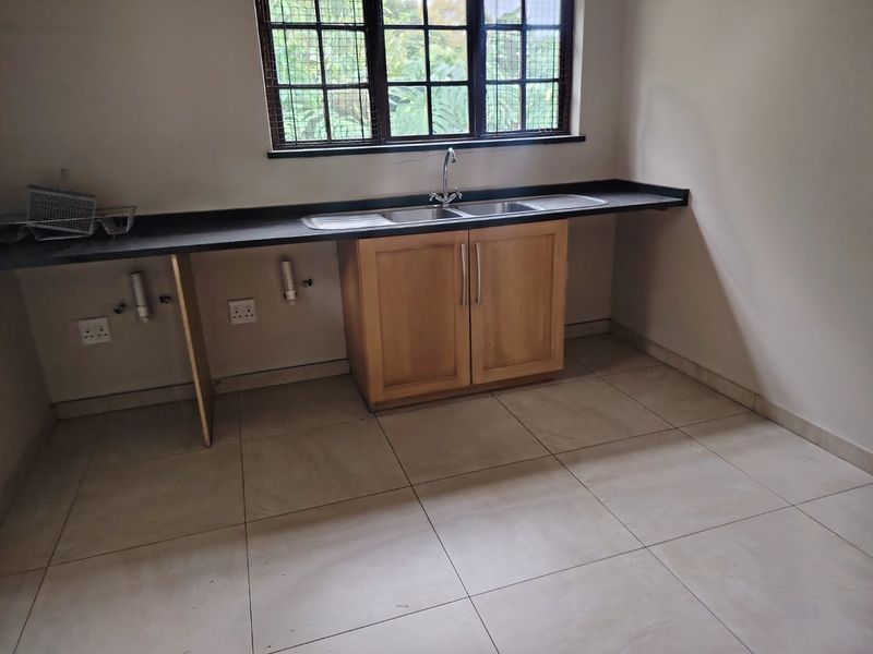 A functional kitchen area featuring a double sink, wooden base cabinetry, dark laminate countertops, and neutral tiled flooring.