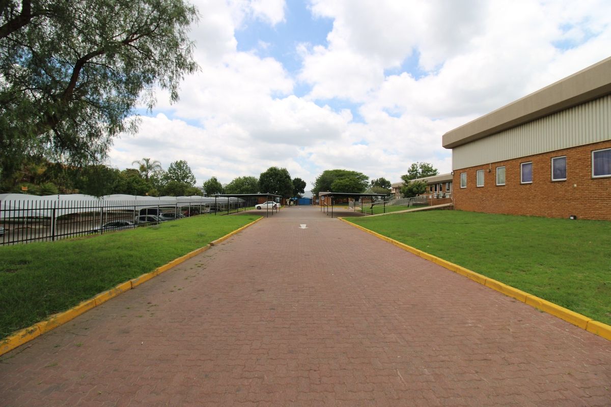 A wide view of the industrial property entrance featuring a paved driveway, covered parking bays, and well-maintained lawn areas surrounding the brick building.