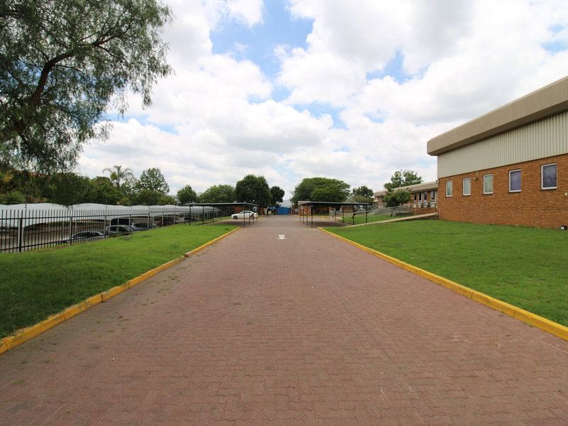 A wide view of the industrial property entrance featuring a paved driveway, covered parking bays, and well-maintained lawn areas surrounding the brick building.