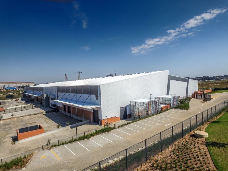 Aerial view of a modern industrial warehouse facility featuring extensive parking, secure perimeter fencing, and large-scale metal cladding construction.