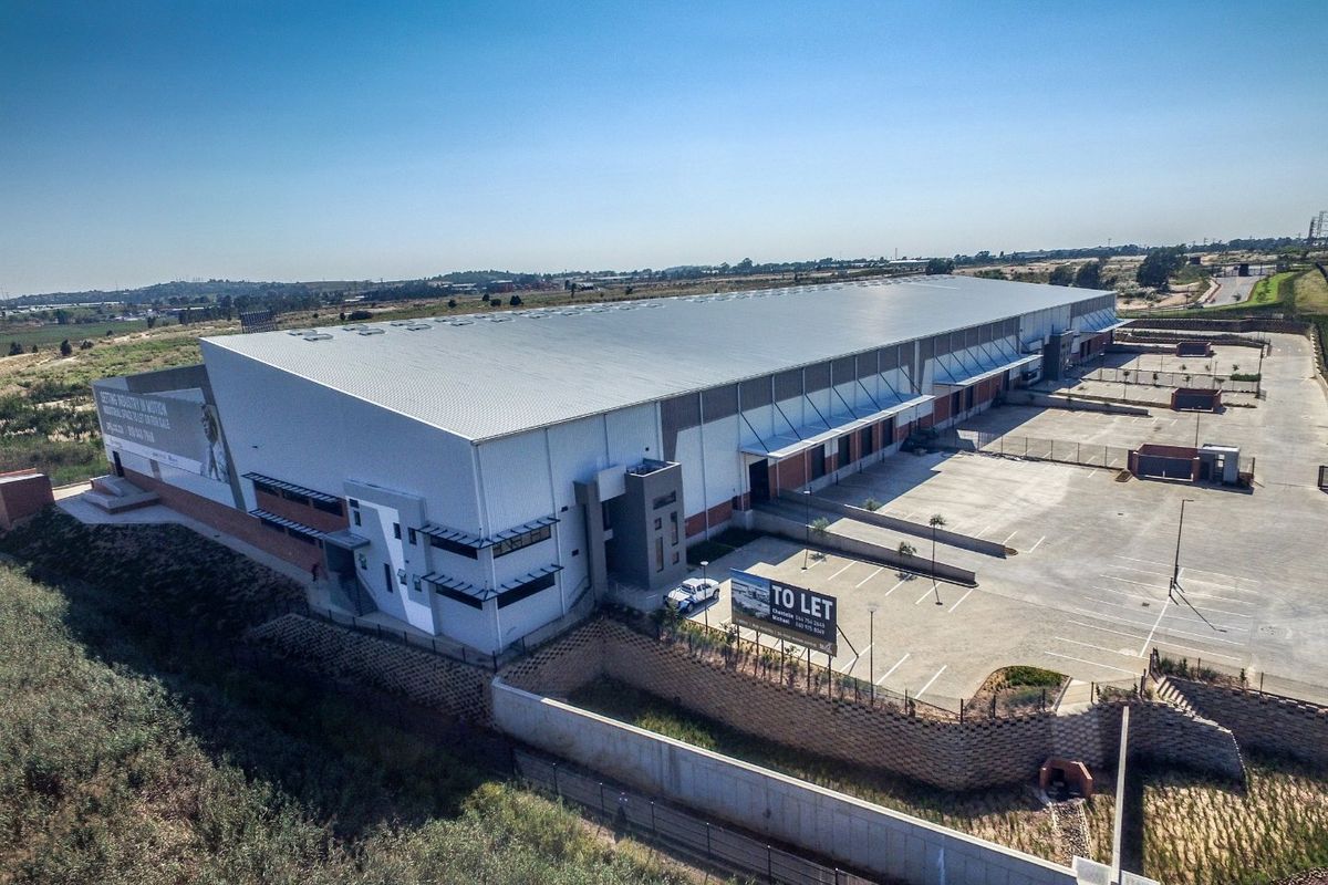 Aerial view of a modern industrial warehouse facility featuring a large metal roof, extensive concrete parking, and secure perimeter fencing.