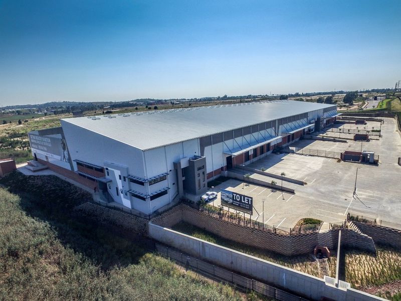 Aerial view of a modern industrial warehouse facility featuring a large metal roof, extensive concrete parking, and secure perimeter fencing.