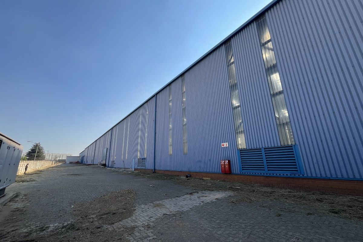 Exterior view of an industrial warehouse featuring corrugated metal cladding, vertical windows, louvered ventilation panels, and a paved yard area.