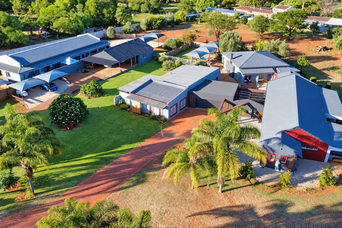 Aerial view of a spacious farm property featuring multiple buildings with metal roofs, extensive landscaped gardens, and a paved driveway.