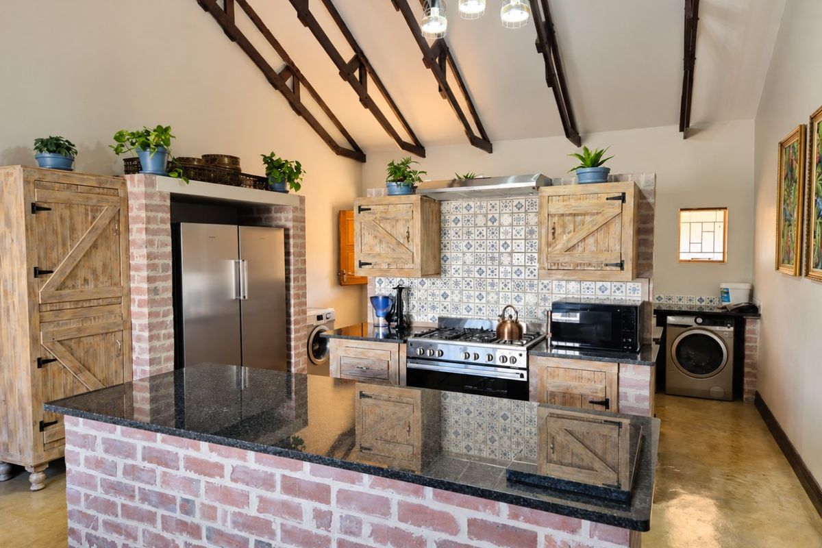 A rustic farmhouse kitchen featuring exposed timber trusses, a brick island with granite countertops, gas stove, and custom wooden cabinetry with decorative tile backsplash.