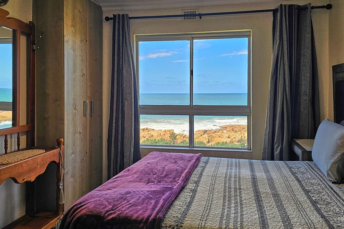 A cozy bedroom featuring a built-in wardrobe and a large window offering a direct, scenic view of the ocean and rocky coastline.