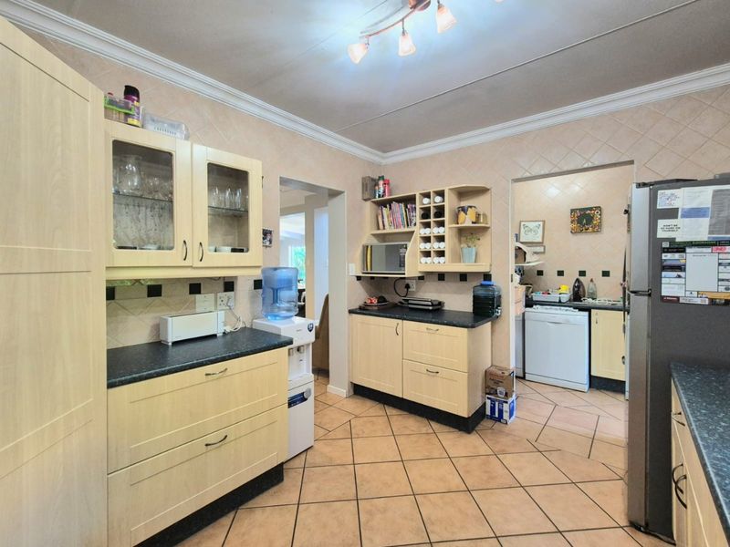 A functional kitchen featuring light-toned cabinetry, dark laminate countertops, and tiled flooring, with an adjacent scullery area visible through the doorway.