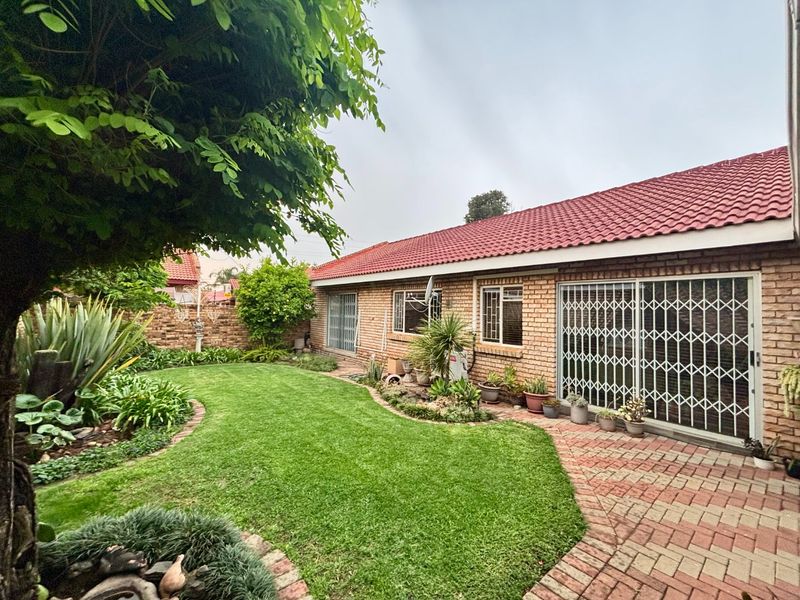 A well-maintained rear exterior view of the townhouse featuring a brick facade, tiled roof, paved patio area, and a landscaped garden with a lawn.